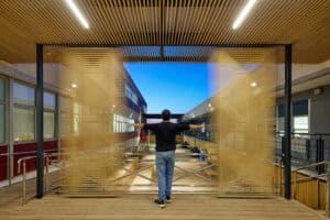 A person stands on a wooden platform, pushing open large slatted במבוק דק gates, revealing a modern building courtyard under a clear sky.