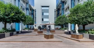 Modern outdoor courtyard with במבוק דק decking, geometric sculptures on pedestals, symmetrical rows of green trees, and a central seating area with umbrellas, flanked by multi-story buildings.