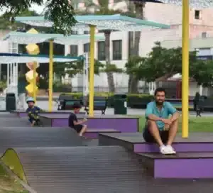 Three people sit on purple steps in an outdoor park area with yellow poles and decorative canopies, surrounded by trees and benches—an inviting setting that could be enhanced with התקנת דק במבוק for added style and comfort.