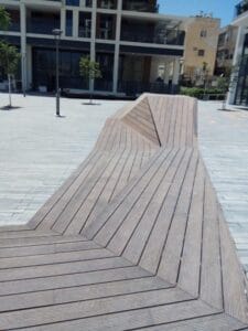 Wooden geometric bench with angular, raised sections sits in an open urban plaza surrounded by modern buildings and a few young trees, featuring במבוק דק for a natural and contemporary touch.
