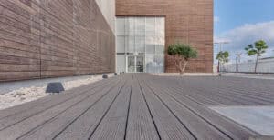 A modern building with wooden paneling and a glass entrance, featuring התקנת דק במבוק, small trees, and white rocks in the landscaped area.