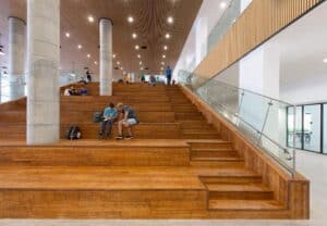 Wide wooden tiered seating area with scattered people, next to glass railings and large columns, in a modern, well-lit building—showcasing inspiration for בניית דקים or incorporating דקים למרפסת in contemporary indoor spaces.