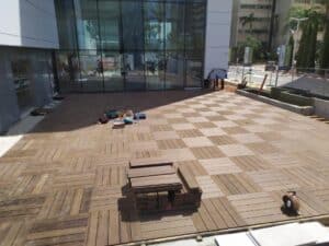 Large outdoor wooden deck under construction outside a modern glass building, with tools and materials scattered on the surface, showcasing התקנת דק במבוק for a stylish and durable addition to your מרפסת.