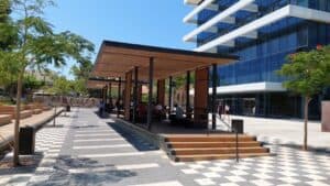Outdoor seating area with people under a modern pergola, next to a glass office building, surrounded by trees and a checkered walkway featuring במבוק דק for an elegant touch.