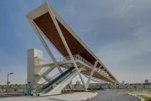 Modern pedestrian bridge with an escalator and stairs crossing a road in an urban area, featuring a large white steel and wood structure with חיפוי במבוק adding a natural touch.