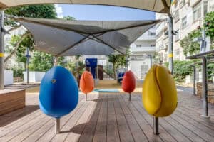 A modern playground with colorful egg-shaped seats on wooden decking ודקים למרפסת, shade canopy above, and residential buildings in the background.