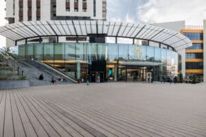 A modern circular glass building with a white overhang, large windows, and people sitting on steps outside on a spacious wooden plaza featuring דקים למרפסת.