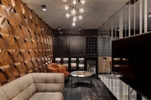 Modern lounge with geometric wooden accent wall featuring חיפוי במבוק, contemporary light fixture, beige and rust seating, round coffee tables, wine storage, and glass partition.