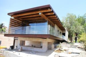 Modern house with exposed wooden beams, glass railings, and stone exterior features בניית דקים למרפסת, elevated above a landscaped yard with rocks and small plants.