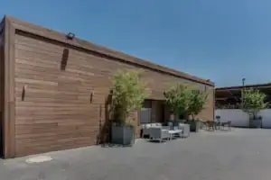 A modern, single-story building with a wooden exterior and במבוק דק, large potted plants, outdoor seating, and a paved courtyard under a clear blue sky.