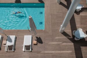 A person swims in a rectangular pool beside a wooden deck with white lounge chairs, closed umbrellas, and round tables. דקים למרפסת and bamboo accents add a stylish, natural touch to the relaxing poolside atmosphere.