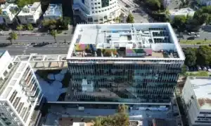 Aerial view of a modern rectangular office building with glass windows, rooftop equipment, and colorful mural sections, featuring spaces for דקים למרפסת or even התקנת דק במבוק, surrounded by roads and other structures.