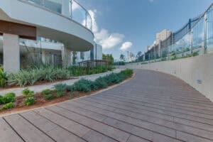 A curved wooden walkway bordered by plants runs alongside a modern building with glass railings, featuring במבוק דק, under a partly cloudy sky.