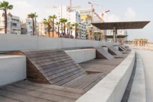 A row of modern במבוק דק and concrete benches lines a seaside promenade with palm trees, city buildings, and construction cranes in the background.