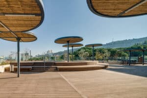Open wooden deck area with circular metal and wood shade structures, including פרגולה במבוק, steps, railing, and a backdrop of trees and distant city buildings under a clear sky.