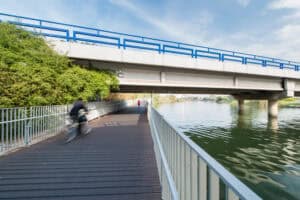 A cyclist and two pedestrians use a במבוק דק wooden path beside a river under a concrete bridge with blue railings, on a clear day.