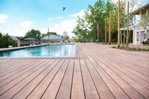 A large wooden deck, perfect for בניית דקים או דקים למרפסת, surrounds a rectangular outdoor swimming pool, with trees, a building, and a bird flying in the sky in the background.