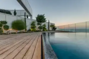 Modern rooftop pool with במבוק דק wooden deck, glass railing, and landscaped plants, overlooking a calm ocean at sunset.