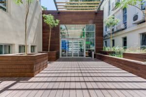 A modern wooden deck walkway, perfect for דקים למרפסת, leads to a glass entrance flanked by large planter boxes with small trees, nestled between two light-colored buildings.
