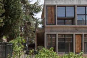 A modern concrete building with large glass windows and vertical wooden panels, surrounded by greenery and trees, features an exterior staircase and stylish דקים למרפסת for a natural touch.