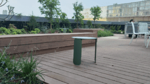 A wooden deck with במבוק דק, a small green metal table, plants, trees, benches, and white chairs; a person sits in the background.
