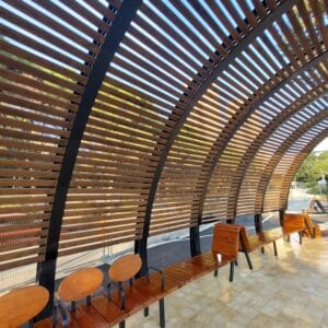 Curved wooden slat structure with built-in benches and chairs, featuring במבוק דק for a natural look, provides a shaded seating area on tiled flooring outdoors.