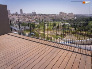 A wooden balcony with a metal railing and פרגולה במבוק overlooks a cityscape with buildings, greenery, and a clear sky in the background.