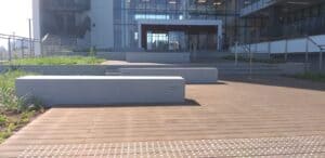 Outdoor view of a modern building entrance featuring במבוק דק decking, concrete benches, stairs, railings, and tactile paving on a sunny day.
