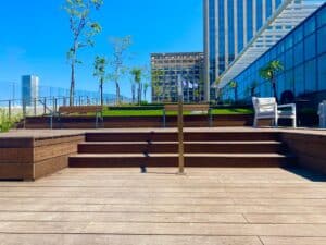 A rooftop deck with wooden steps, a handrail, seating area, small trees, and skyscrapers in the background under a clear blue sky—enhanced by התקנת דק במבוק for a stylish and sustainable outdoor retreat.