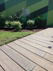 Wooden walkway next to a patch of grass and small bushes, with a green wall in the background displaying the "Park Bavli" logo—an ideal spot להתקנת דק במבוק or exploring דקים למרפסת for a natural look.