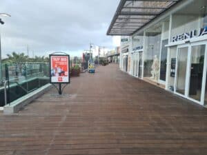 Outdoor shopping promenade with בניית דקים wooden flooring, store fronts to the right, glass railing on the left, and an advertisement display; area appears mostly empty.