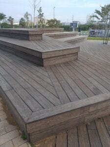 Multiple wooden platforms and steps, including במבוק דק, arranged in geometric patterns in an outdoor park setting with trees and distant buildings visible.