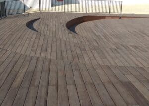 Wooden deck with two curved sunken seating areas and metal railing in the background; sunlight casts shadows on the surface, highlighting איכות בניית דקים and modern דקים למרפסת design.