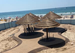 Wooden boardwalk with three thatched sun shelters on a sandy beach, featuring בניית דקים craftsmanship. People and the ocean are visible in the background.