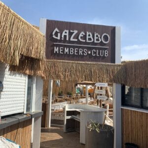 Entrance to a thatched-roof outdoor area with a sign reading "Gazebbo Members Club," bamboo cladding (חיפוי במבוק), and directional signs for shower, lounge, and beach.