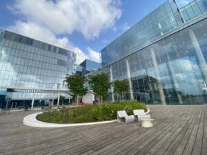 Modern office building with glass facades, outdoor seating on במבוק דק wooden decking, and a small landscaped garden with trees and plants under a partly cloudy sky.