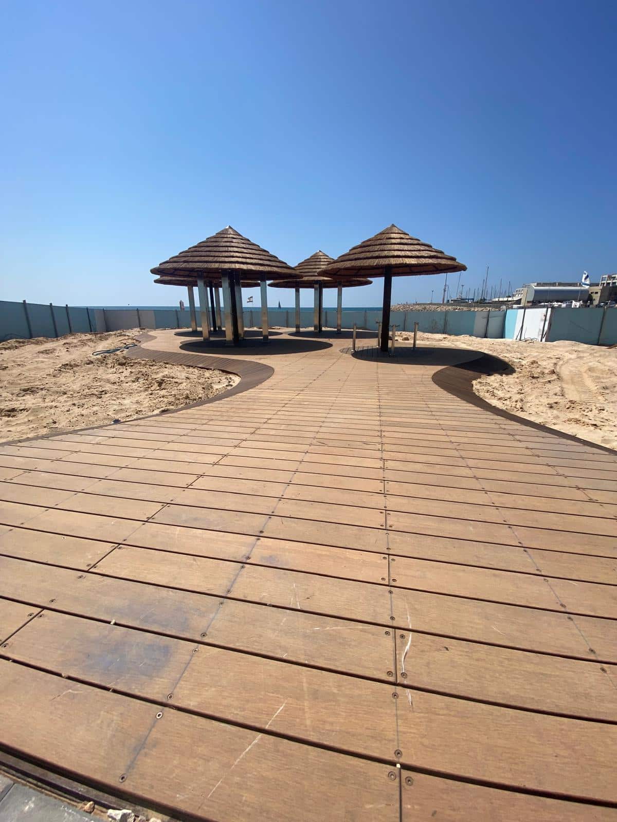 Wooden boardwalk leading to shaded huts on a sandy area, featuring התקנת דק במבוק and פרגולה במבוק, with blue fencing and a clear sky in the background.