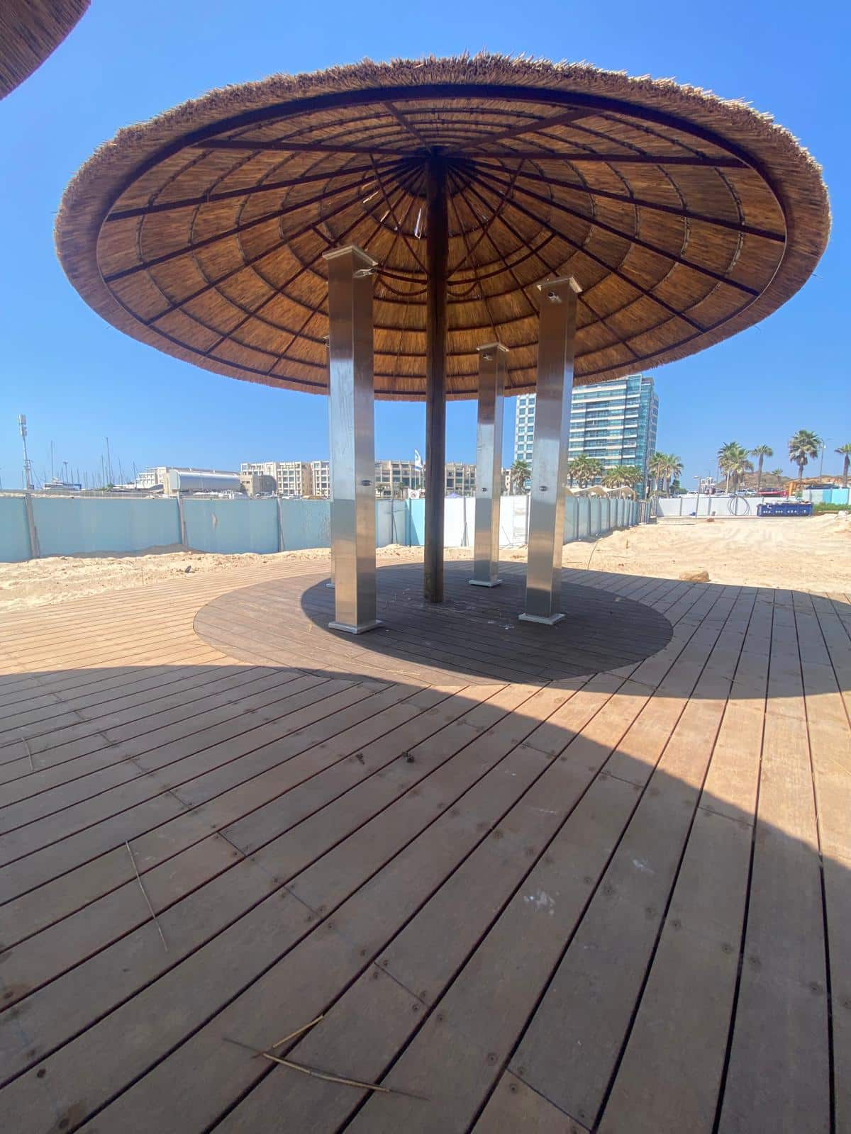 Large circular wooden and steel shade structure, resembling a פרגולה במבוק, on a boardwalk with a building, palm trees, and clear blue sky in the background.