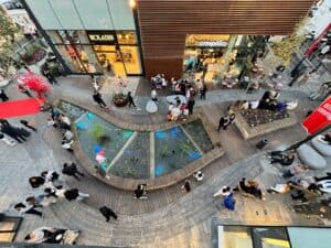 Aerial view of a modern shopping area with people walking, a small pond with plants, storefronts like Roladin and Crespella, and stylish פרגולה במבוק shading the walkway.